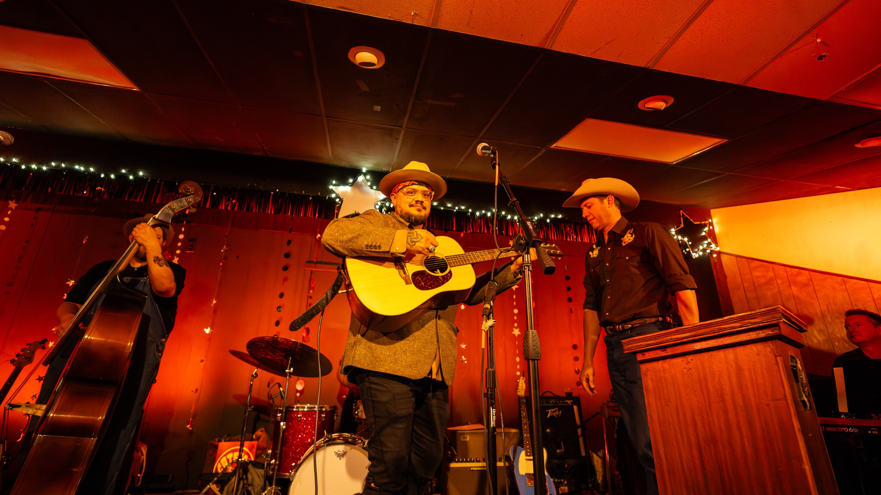 A man strums a guitar onstage backed by a band lit up in a bright honky tonk red.