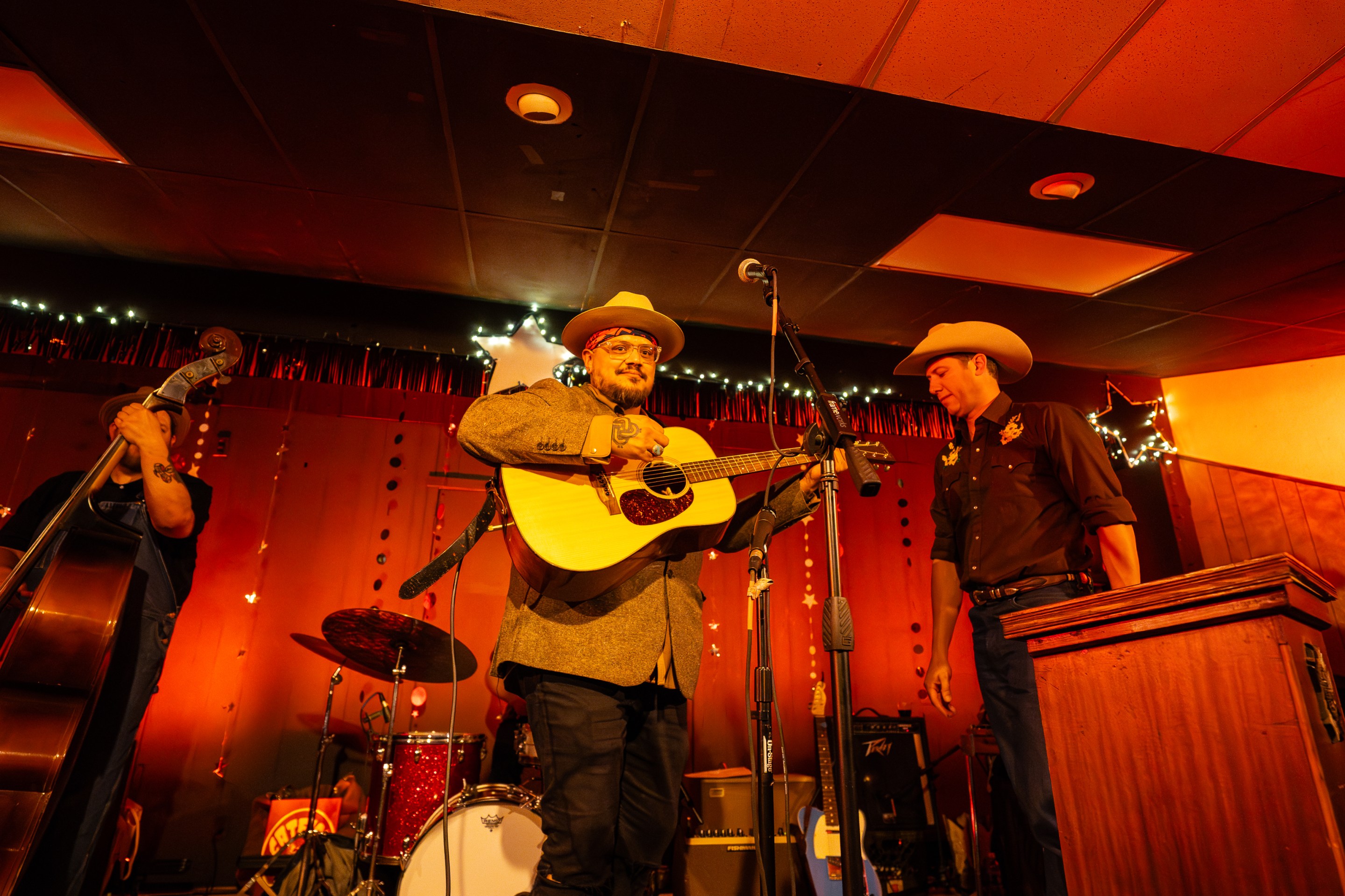 A man strums a guitar onstage backed by a band lit up in a bright honky tonk red.