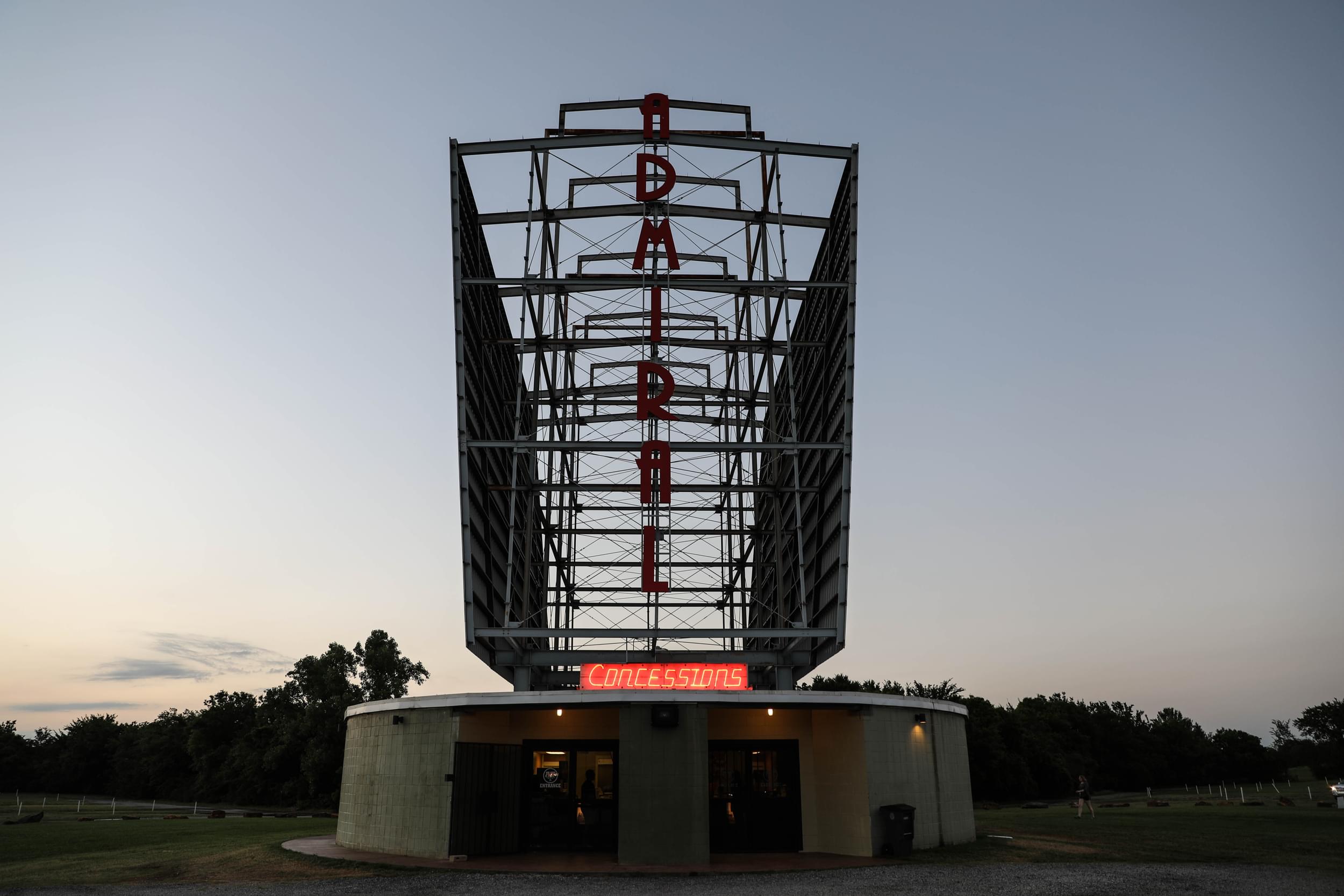 A neon light concession stand shines underneath a nine-story-high drive-in movie theater screen.