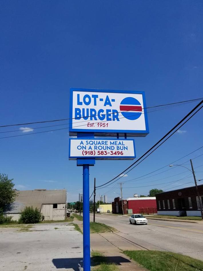 Blue and red sign reading "Lot-A-Burger" in front of blue sky and parking lot.