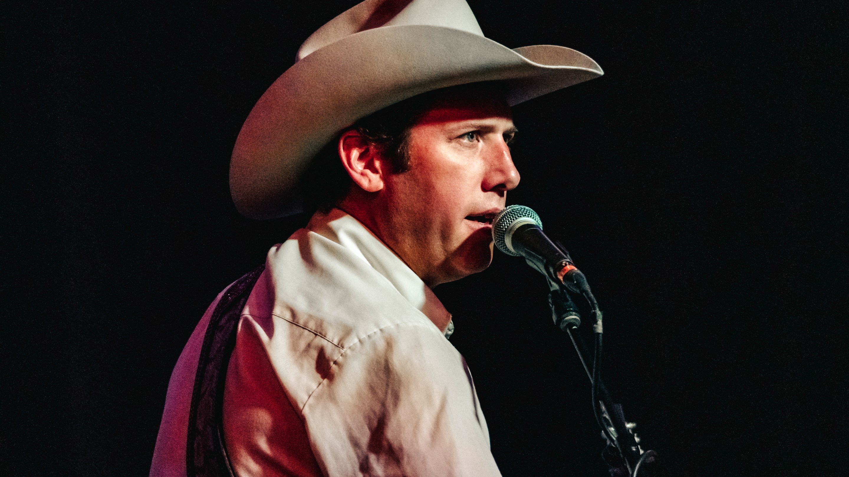 A man in a cowboy hat sings into a microphone while staring straight ahead into the stage lights.