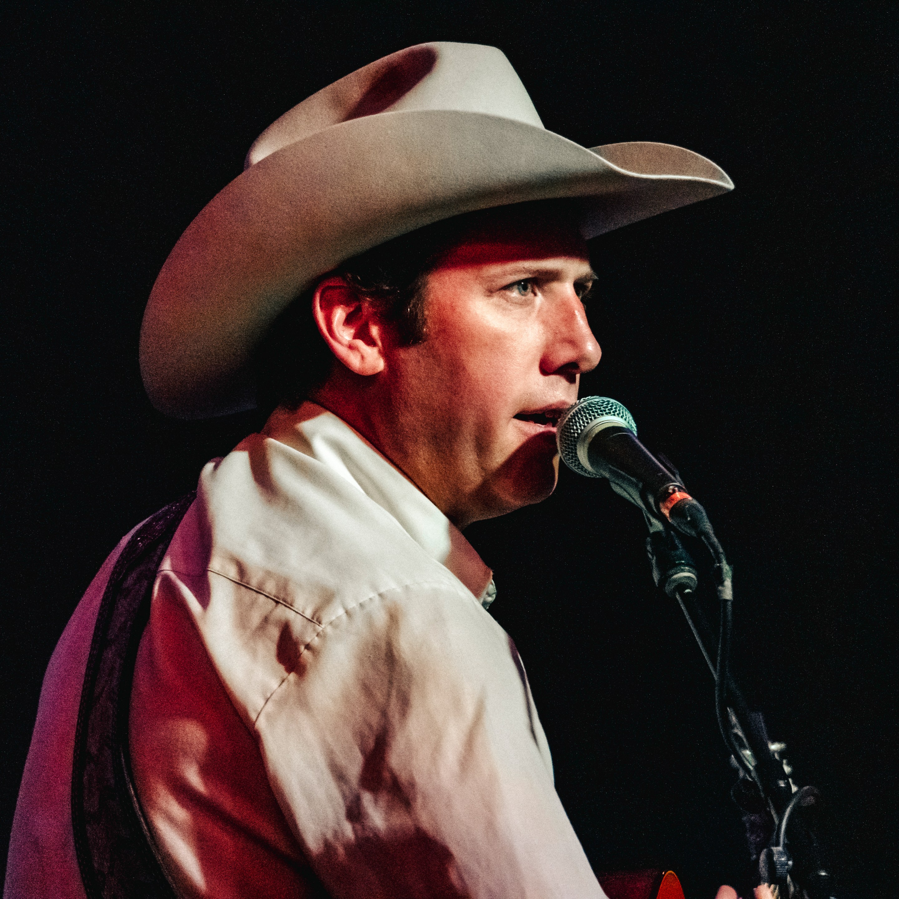 A man in a cowboy hat sings into a microphone while staring straight ahead into the stage lights.
