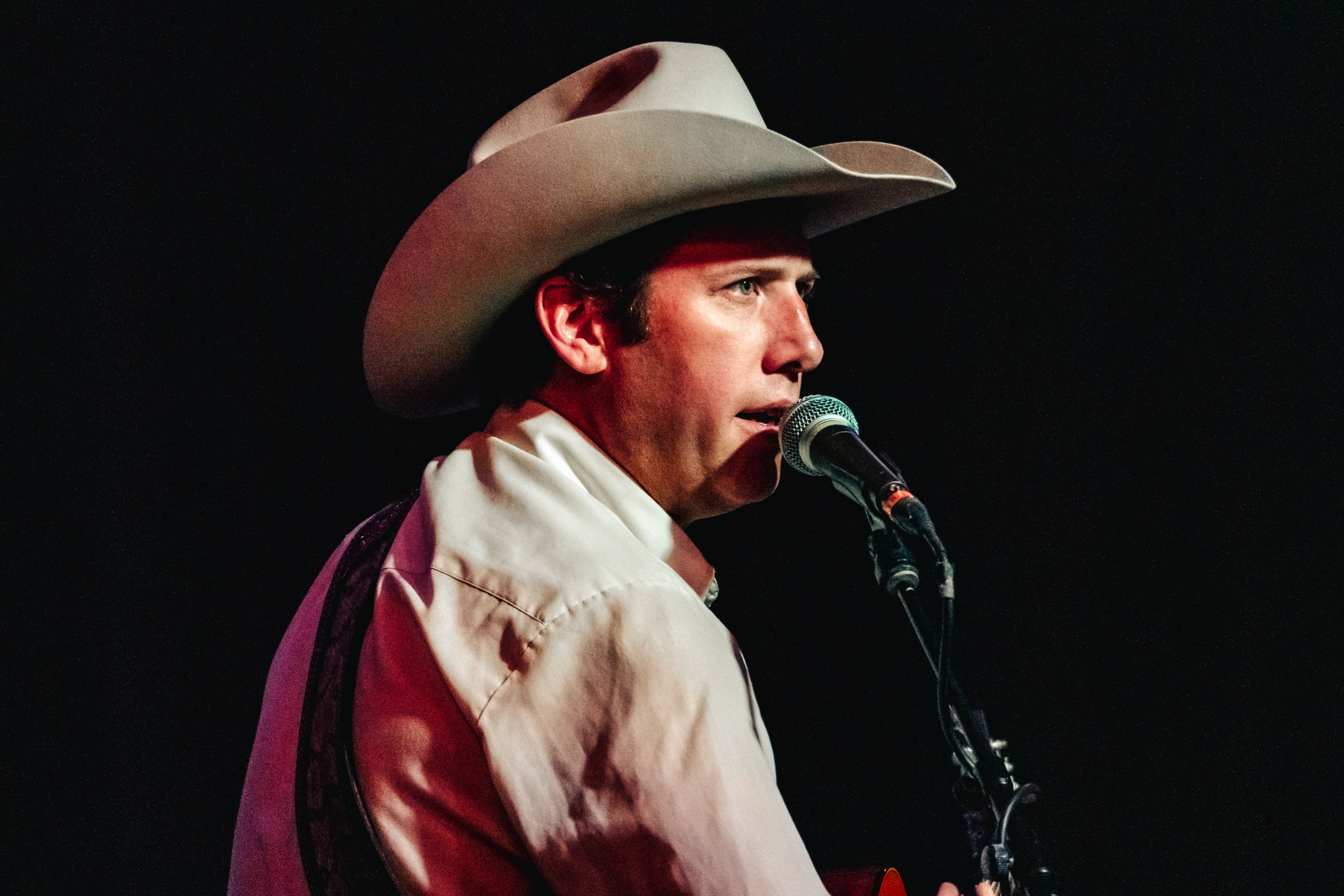 A man in a cowboy hat sings into a microphone while staring straight ahead into the stage lights.