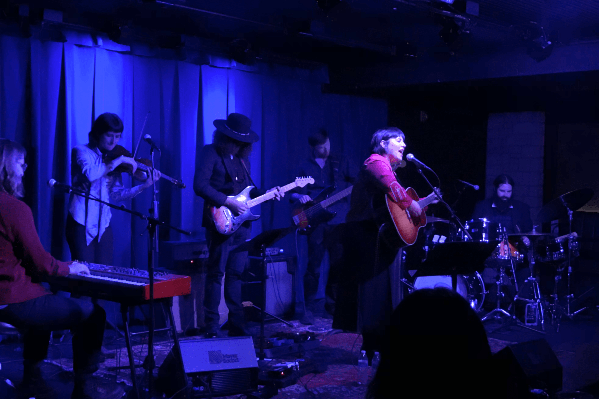 A woman, under a spotlight, plays a guitar and sings onstage in a dimly lit speakeasy on a stage.