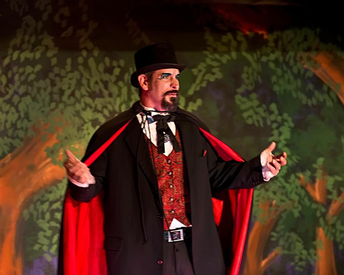 An actor stands in a suit and cape with their arms extended.