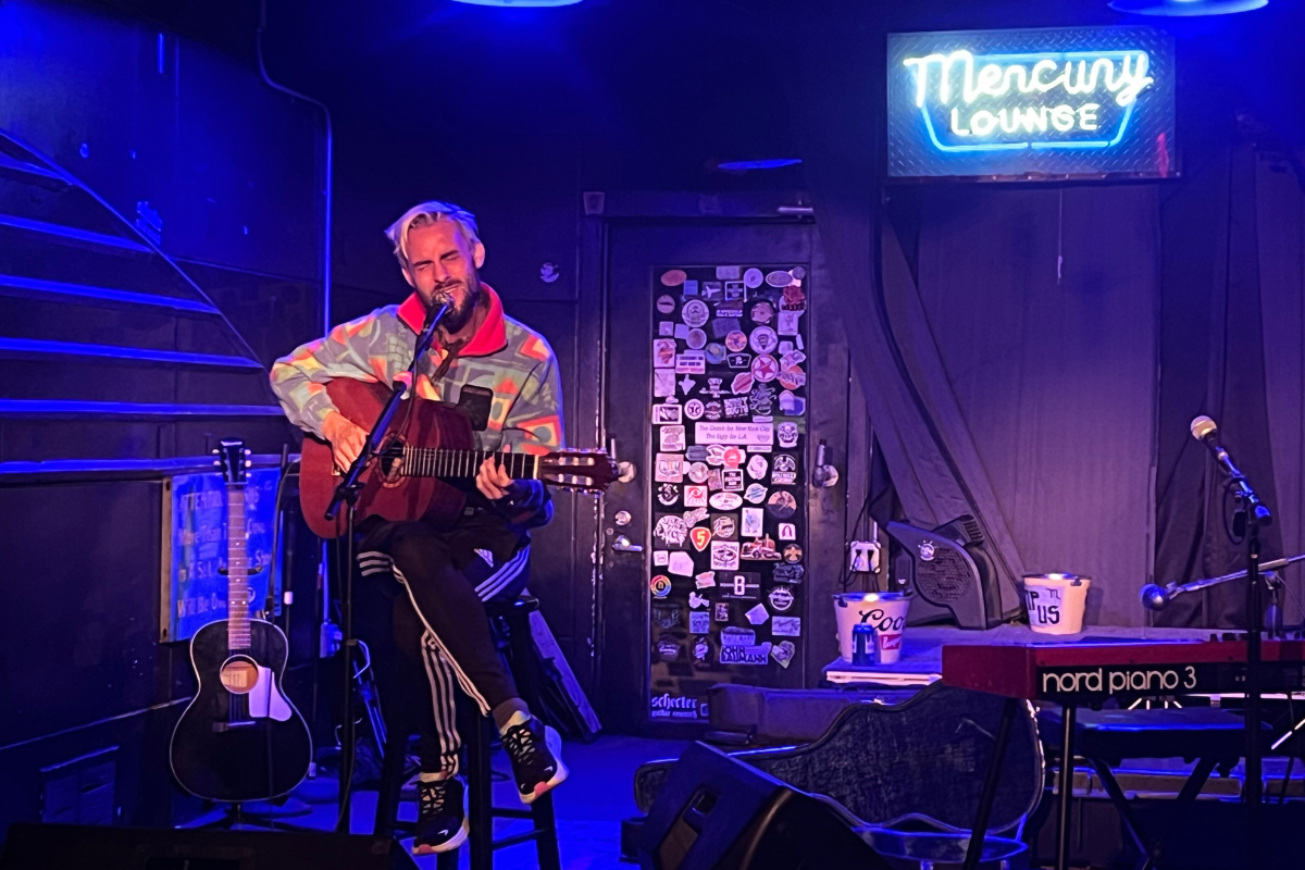 Robert Ellis plays an acoustic guitar and sings on stage in front of the neon Mercury Lounge sign.